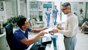 Male healthcare worker in scrubs sits at computer and helps male patient standing at desk