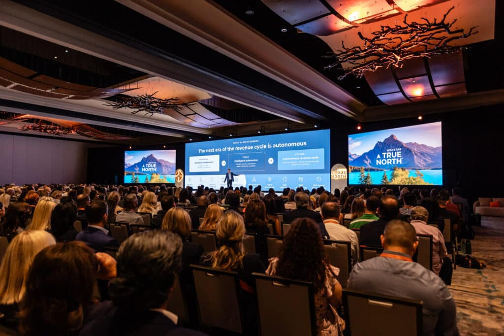Room with large audience and Matt Hawkins, Waystar CEO, on stage in front of 3 large screens.